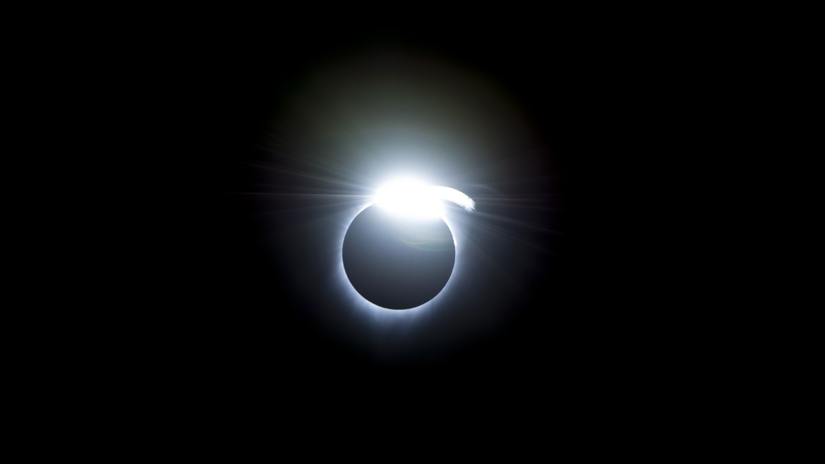 Fotografia de um eclipse solar total no instante do anel de diamante: o disco da Lua completamente escuro recortado sobre o ceu preto, com um unico ponto de luz intensa na borda superior e um halo tenue de coroa em volta