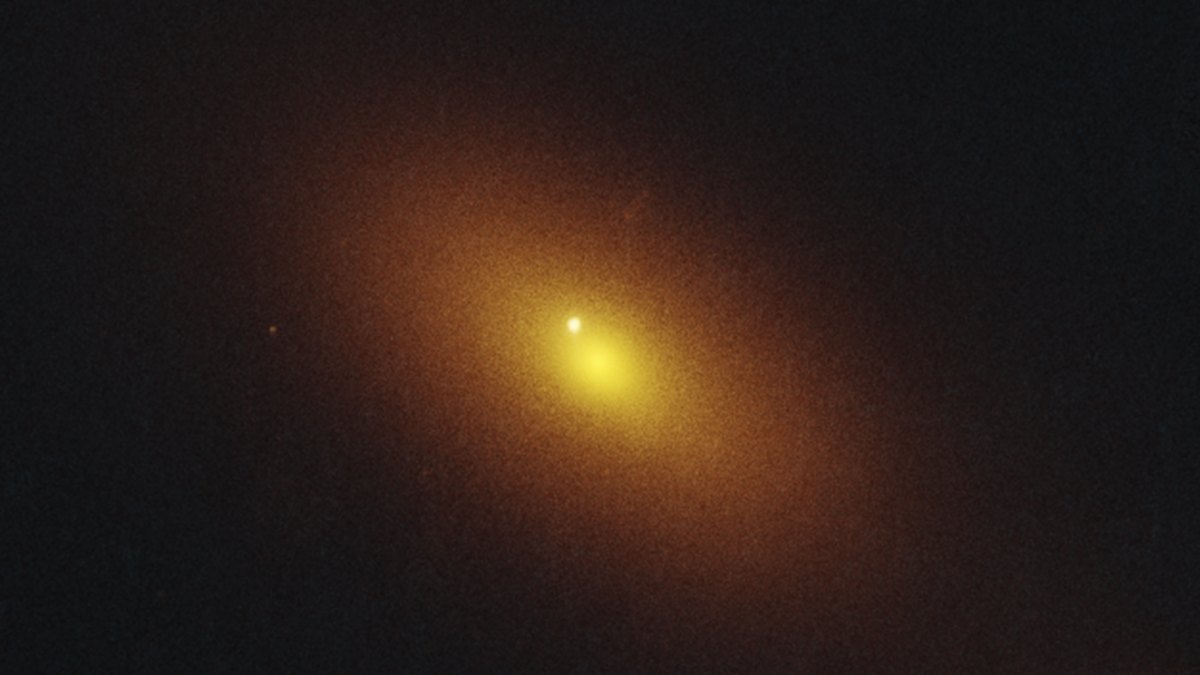 Fotografia do Telescópio Espacial Hubble: uma galáxia de halo alaranjado difuso com núcleo amarélo brilhante e, deslocado dele para cima e para a esquerda, um ponto branco-azulado nítido e isolado, o clarão do evento de disrupção de maré