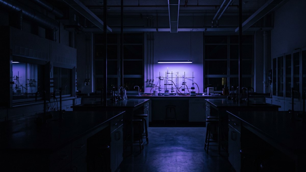 Laboratório de química às escuras, visto de longe: uma única lâmpada estreita sobre uma bancada distante lança luz violeta que recorta silhuetas de vidrarias e suportes metálicos, enquanto o resto da sala se dissolve em penumbra fria